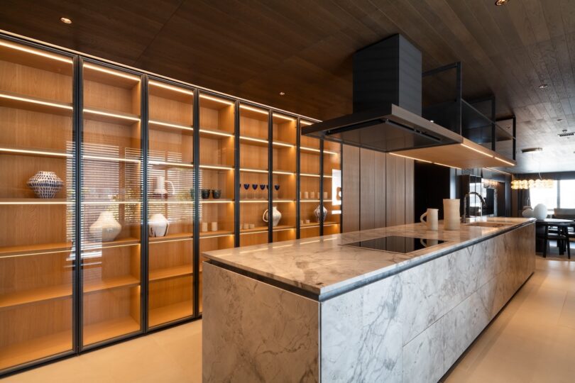 Modern kitchen with a large marble island, sleek cabinet with shelves displaying vases, and wooden ceiling with recessed lighting.