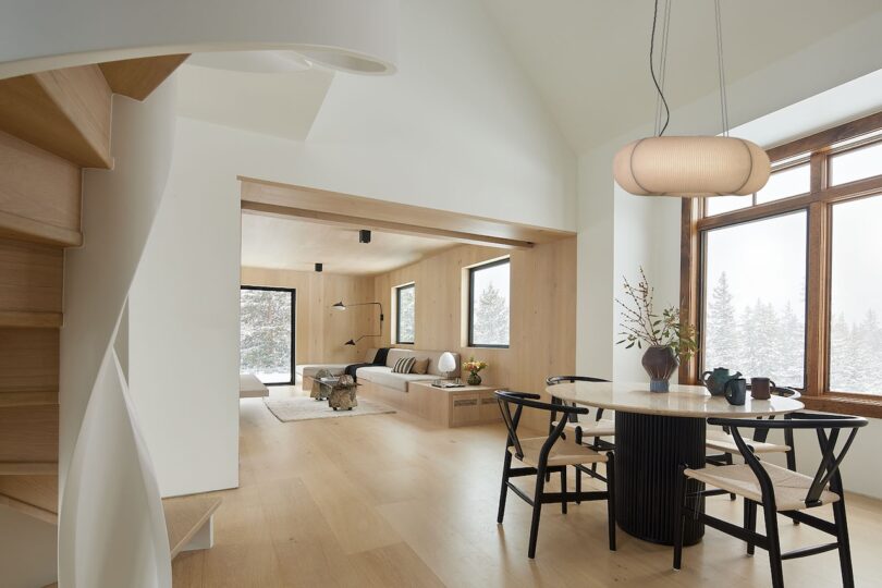Modern interior with light wood floors, a dining area with a round table and chairs, a cozy sunken living area, and large windows showing a snowy landscape outside.
