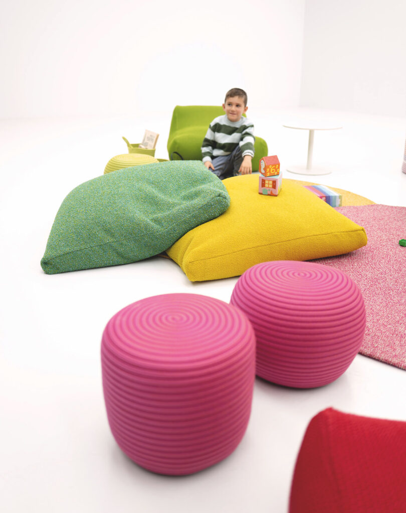 A child sits among colorful cushions and stools in a bright, minimalist room, with toys scattered around