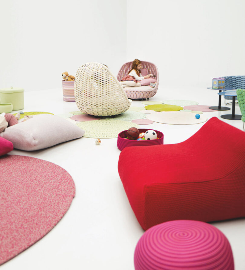 A minimalist room with colorful cushions, a wicker chair, toys, and a child sitting amidst light pink, green, and red decor