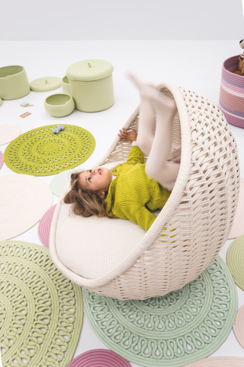 Child in a bright green sweater playfully upside down in a woven chair surrounded by colorful mats and baskets in a light, airy room