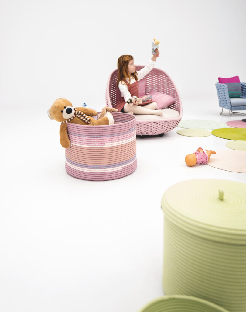 A child sits on a circular pink chair in a white room, holding a toy. A teddy bear is on a striped ottoman nearby, with a doll on the floor next to round rugs.