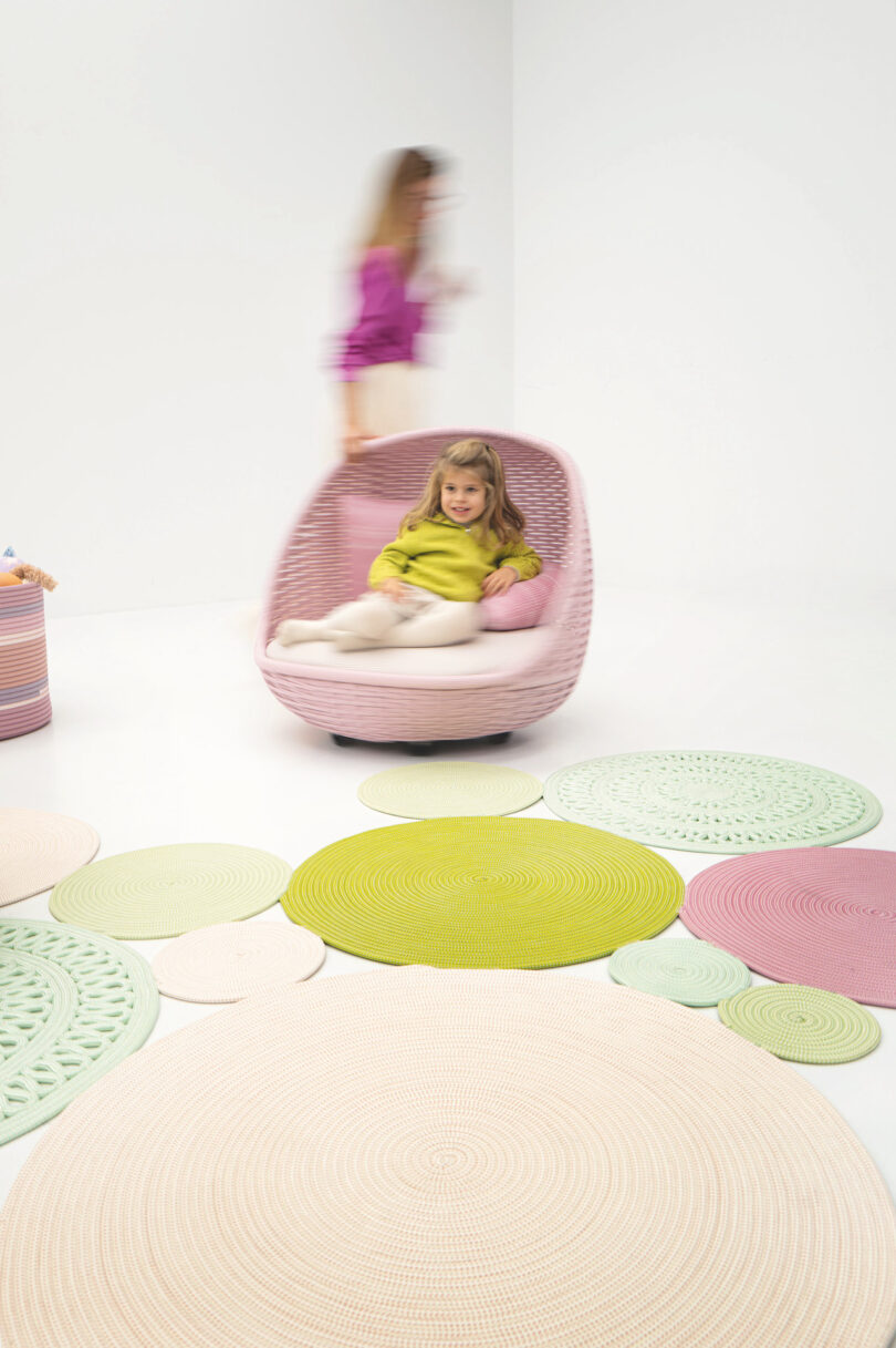 Child sitting in a pink chair in a white room with circular mats on the floor; figure in motion in the background