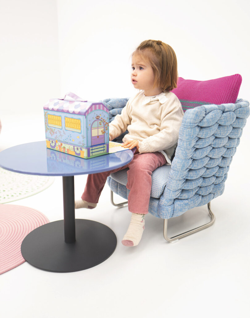 A toddler sits in a blue woven chair, looking at a colorful toy house on a round table, with pink cushions and a patterned rug nearby