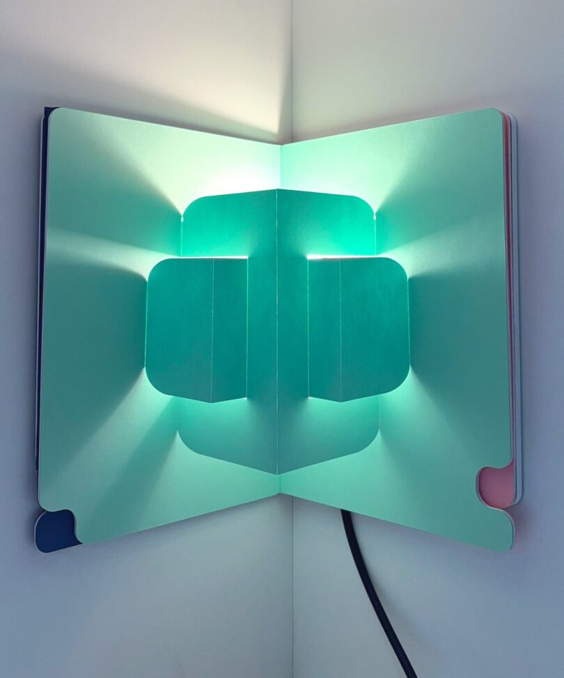 Open book with geometric cutouts and blue-green light casts shadows and glows. The book is placed in the corner of a room, with a visible black cord