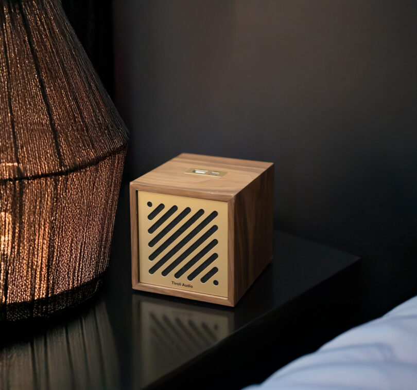 A Tivoli wooden cube-shaped speaker with diagonal slits on the front rests elegantly on a black surface beside a lamp.