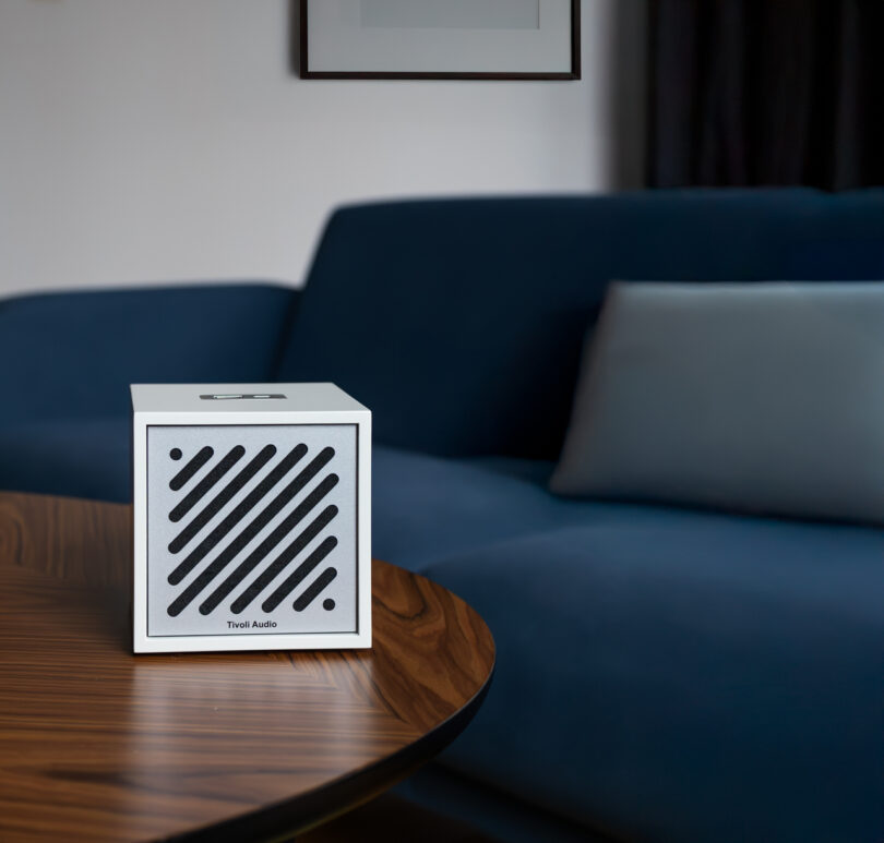 A small Tivoli speaker with diagonal black slats sits on a wooden table in front of a blue couch.