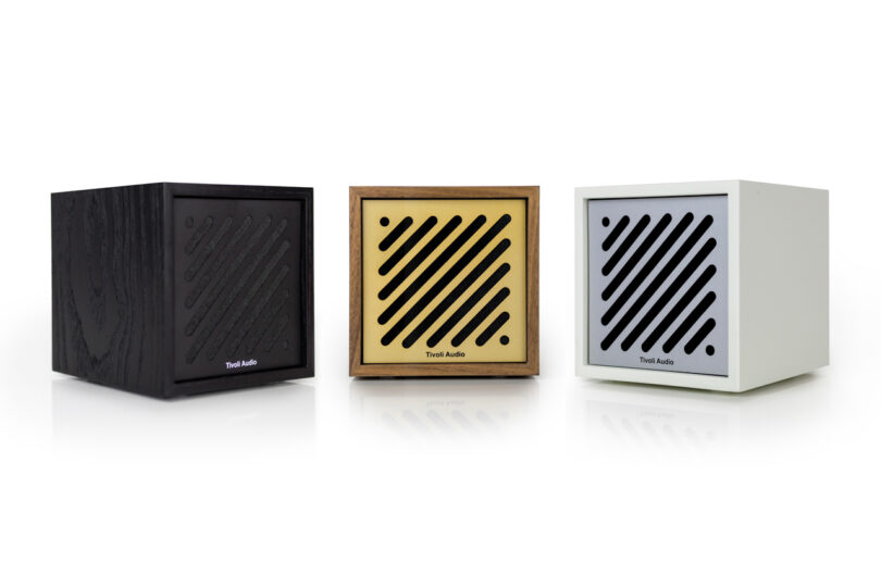 Three cube-shaped Tivoli speakers in black, wood, and white finishes are displayed in a row against a white background. Each features a front grill with diagonal slats and "Tivoli Audio" branding.