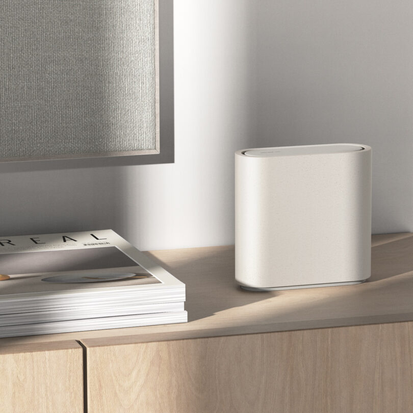 A white, modern wireless router on a light wood cabinet next to a stack of magazines, with part of a framed artwork visible in the background.
