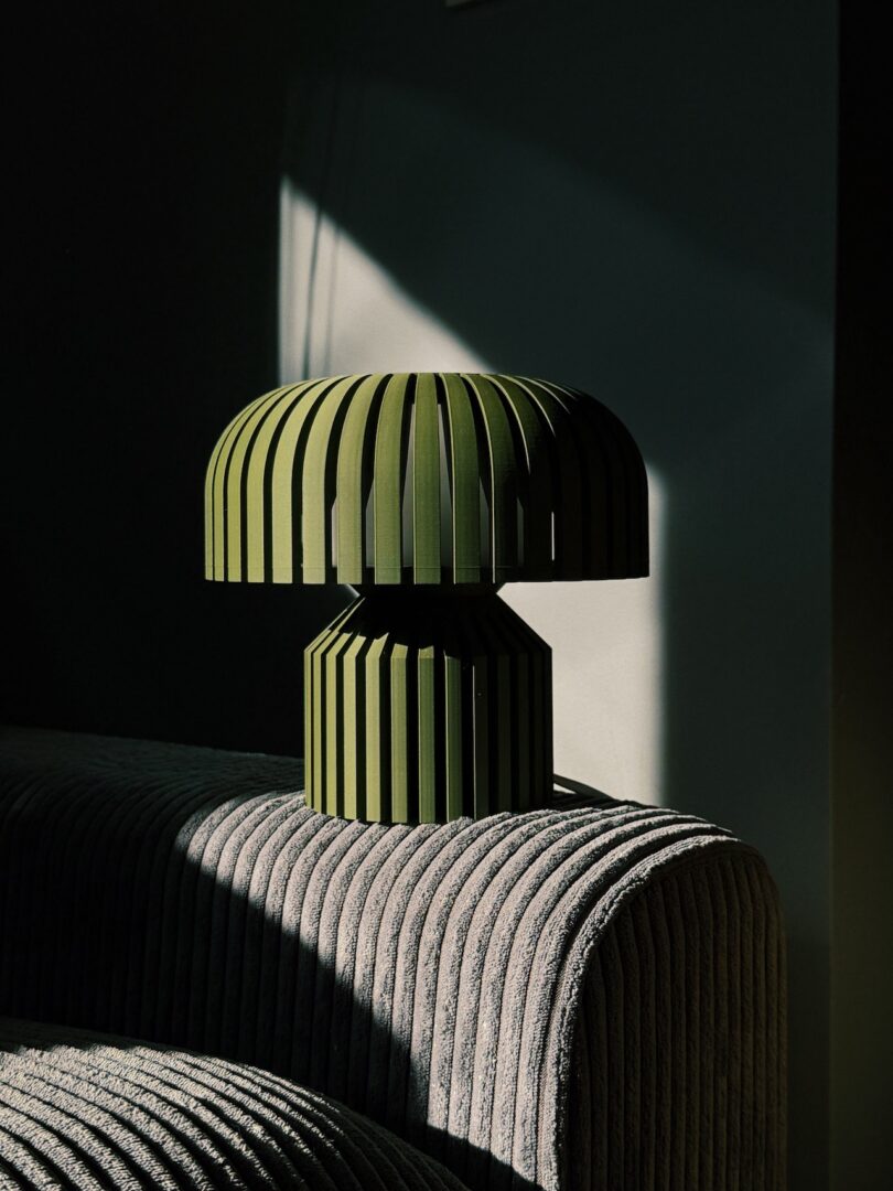 The vibrant Walmen green pleated lamp, with its rounded top, rests on a ribbed fabric surface. Slanting sunlight illuminates it, casting intricate shadows across the dark Dumaliang green wall