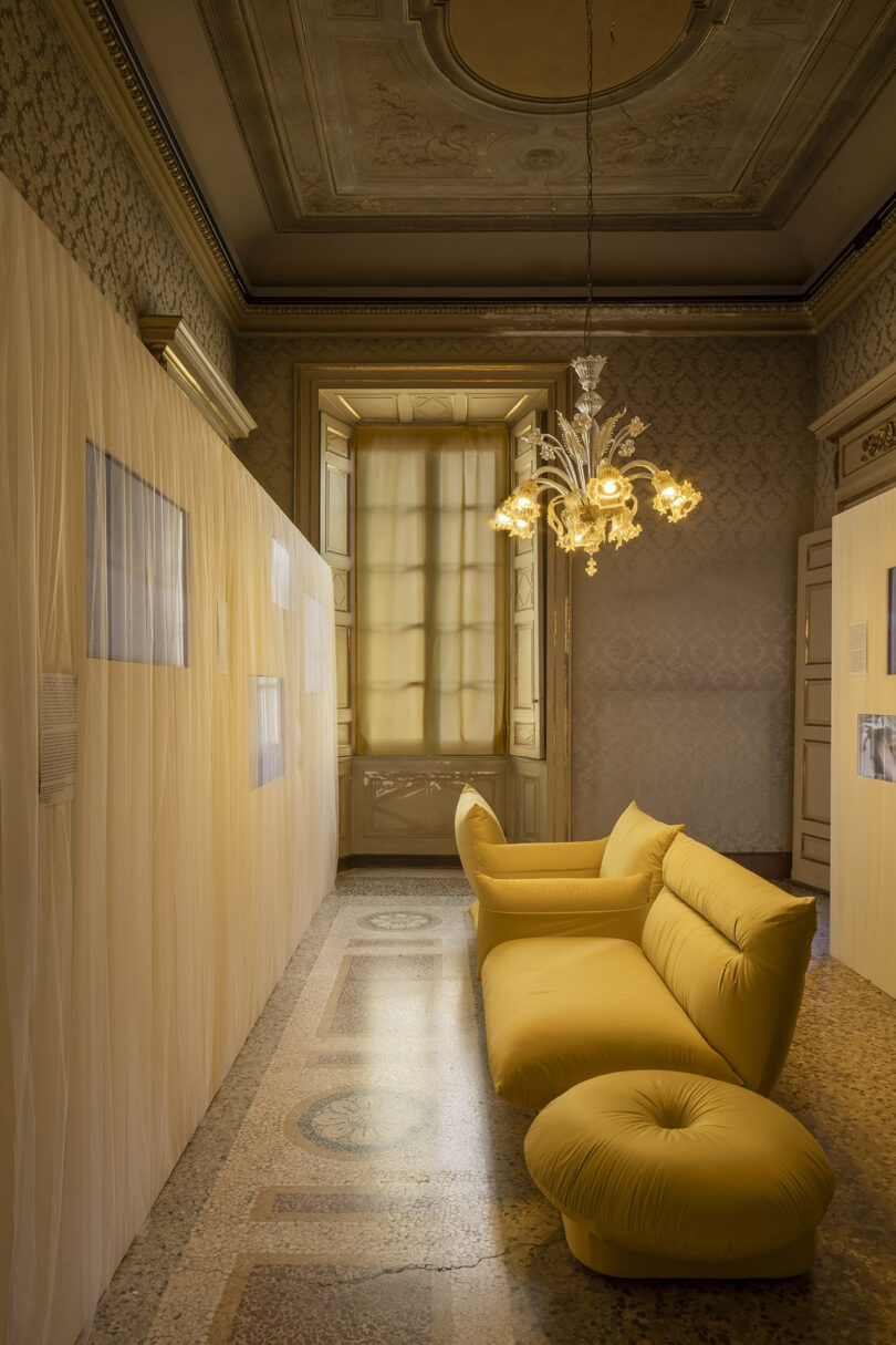 A luxurious room with ornate ceiling, patterned walls, a chandelier, sheer curtains, and modern yellow sofa seating on a decorative stone floor.