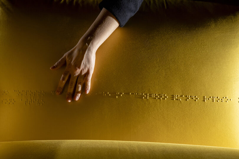 A hand touches a yellow surface with a line of raised Braille dots.