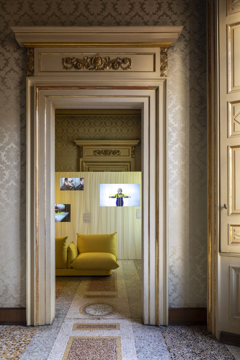 View through ornate doorways into a room with yellow cushioned seating, patterned walls, and illuminated photo displays above the sofa.