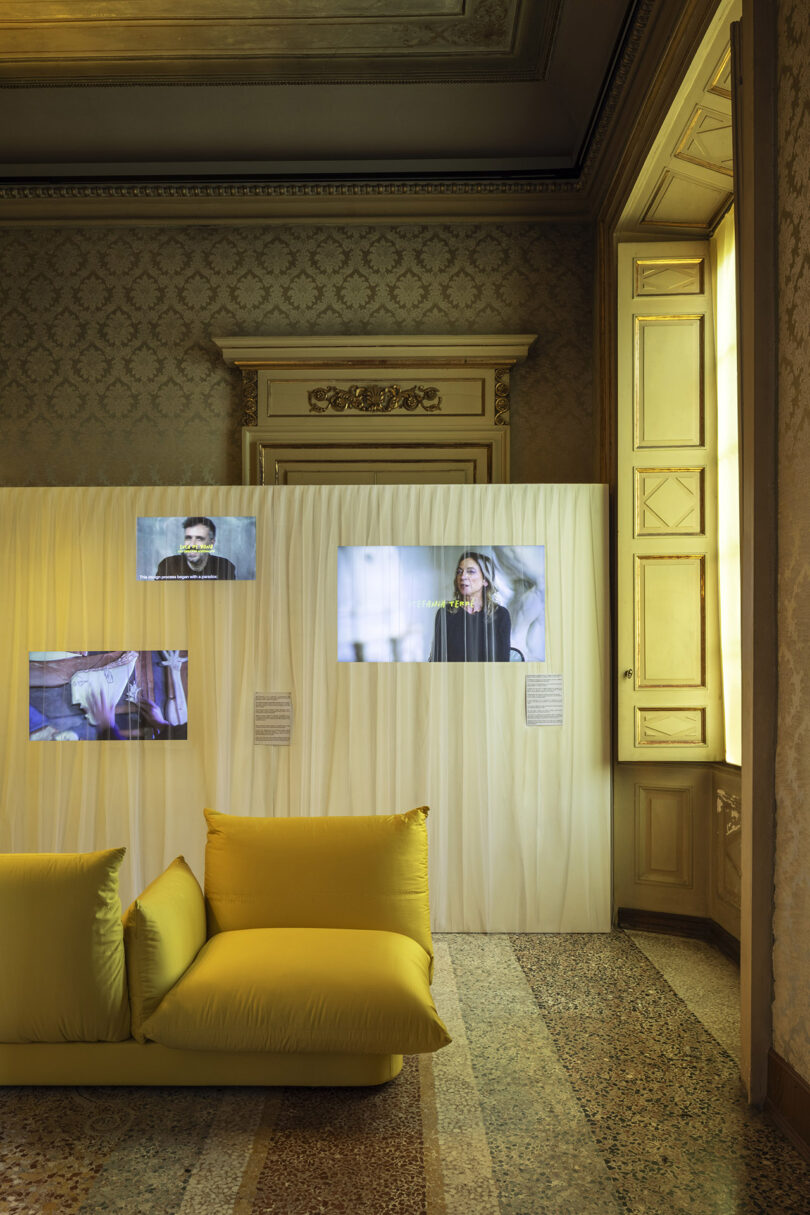 A softly lit room with patterned wallpaper, a yellow cushioned sofa, and a partition displaying three video screens showing people and text. Natural light comes in through a tall window.