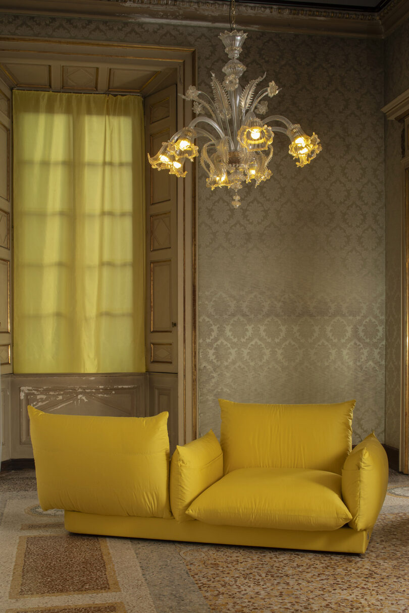 A plush yellow sofa with asymmetrical backrests sits under an ornate glass chandelier in a vintage room with patterned wallpaper and tall windows covered by yellow curtains.