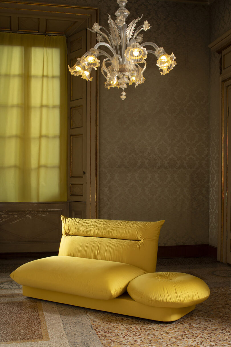 A bright yellow modern sofa sits in a vintage room with ornate wallpaper, mosaic flooring, and a glass chandelier hanging from the ceiling.