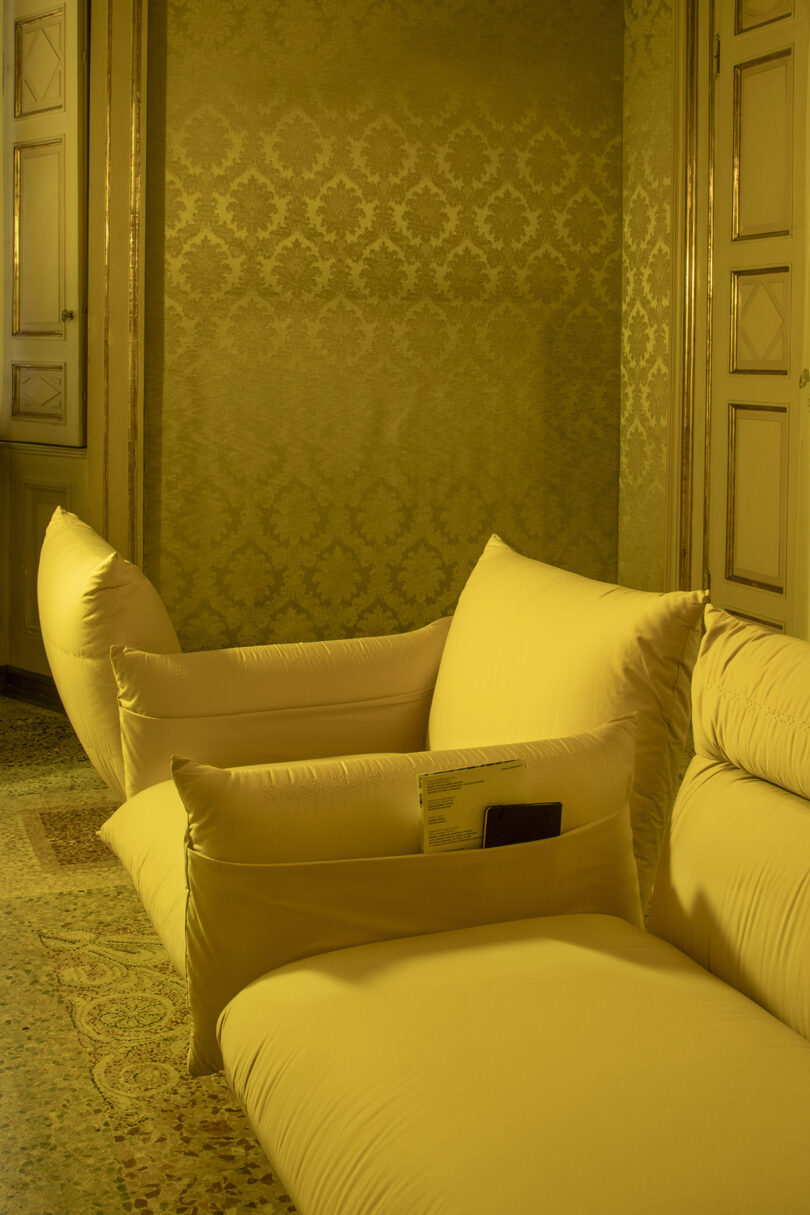 A yellow cushioned sofa with a phone and a small booklet in its side pocket, set in a room with patterned wallpaper and pale wooden doors.