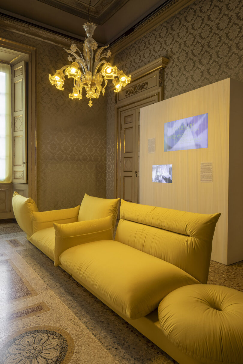 A yellow modern sofa sits in an ornate room with patterned walls, a chandelier, and a display screen mounted on a wooden panel.