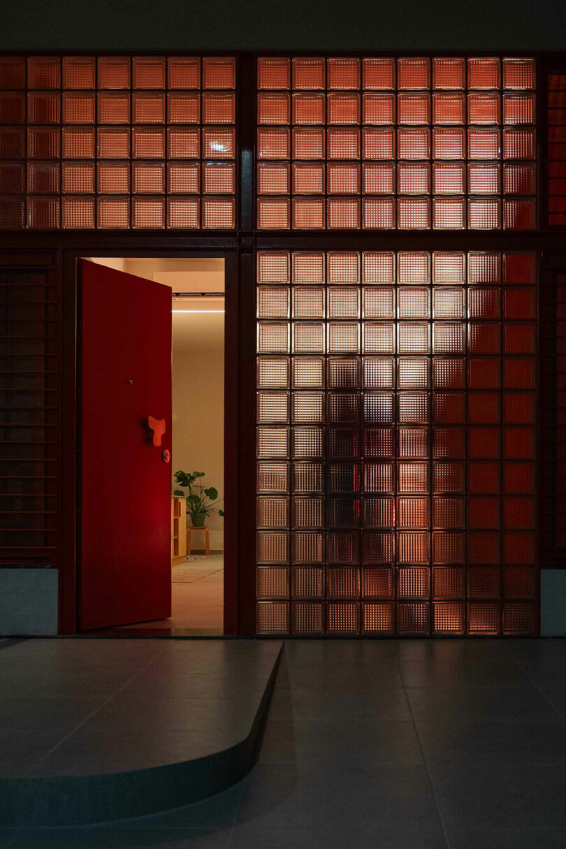 A red door slightly ajar, set in a wall of glass blocks, reveals a softly lit interior with a potted plant visible inside.