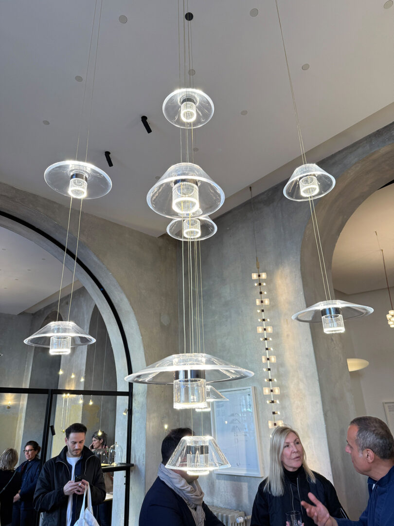 Modern Aura Snøhetta glass pendant lights hang from the ceiling in a stylish, high-ceilinged room with several people standing and talking below.