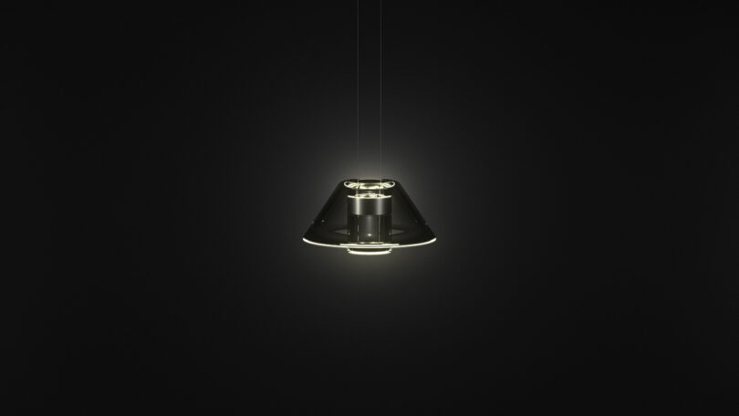 A modern, conical Aura pendant light fixture by Snøhetta emits a soft glow in a dark, minimalist space.