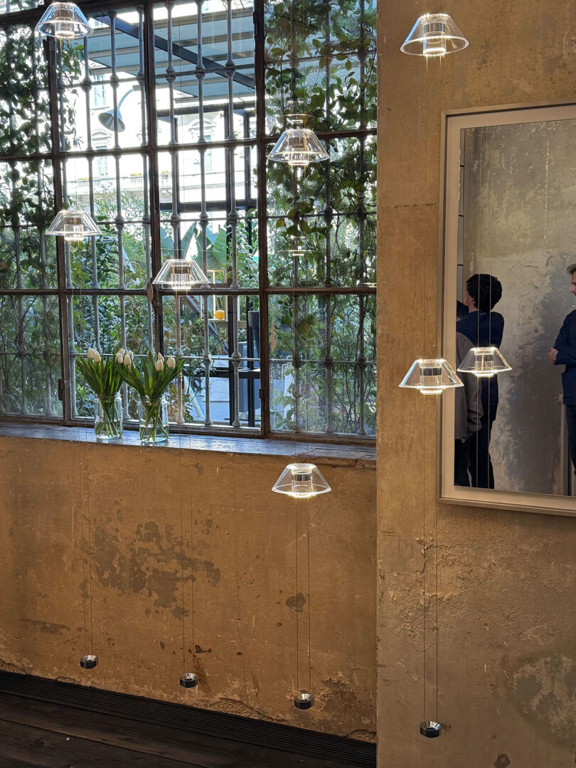 A group of Aura Snøhetta pendant lights hang in front of a wall with large windows and green plants outside. A mirror reflects two people standing nearby.