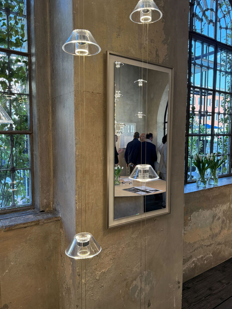 A mirror on a textured wall reflects people standing and talking; pendant lights, Aura Snøhetta-inspired touches, and large windows with green plants are visible in the room.