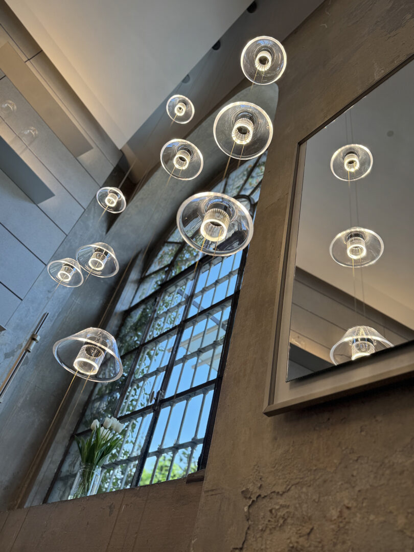 Modern Aura lights from Snøhetta hang from a high ceiling near a large industrial window, reflected in a wall mirror; a vase with white flowers sits below the window.