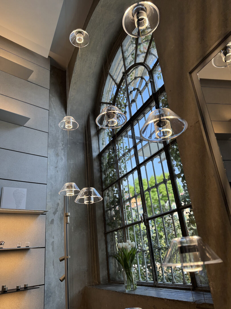 Pendant lights from the Aura collection by Snøhetta hang near a large arched window with black framing, letting in natural light. A vase of white tulips sits on the windowsill.