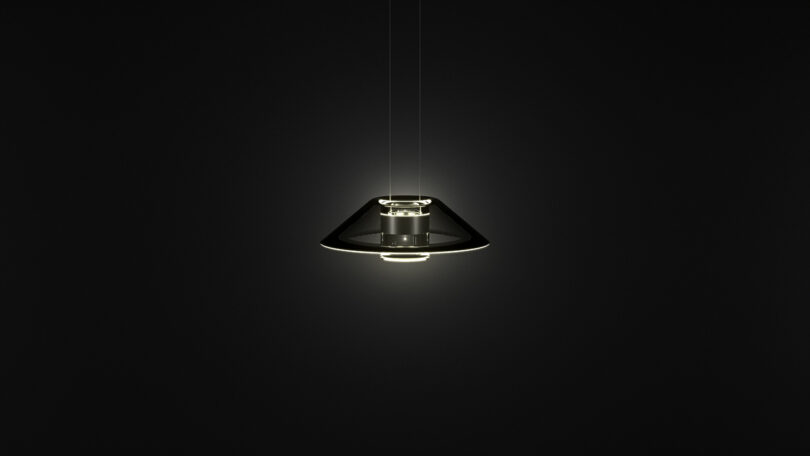 A modern Aura Snøhetta pendant light with a conical glass shade hangs from the ceiling, glowing softly against a dark background.