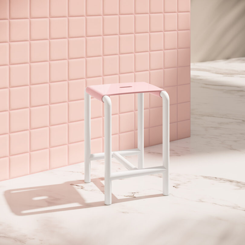 A pink and white stool stands on a marble floor next to a pink tiled wall, with sunlight casting soft shadows.