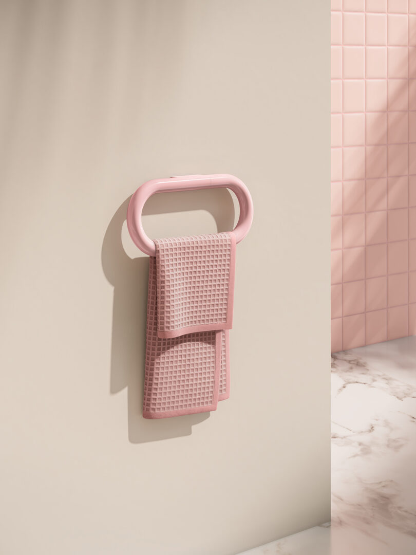 A pink towel is neatly folded and hung on a pink oval towel holder mounted on a beige wall, with pink tiled and marble surfaces in the background.