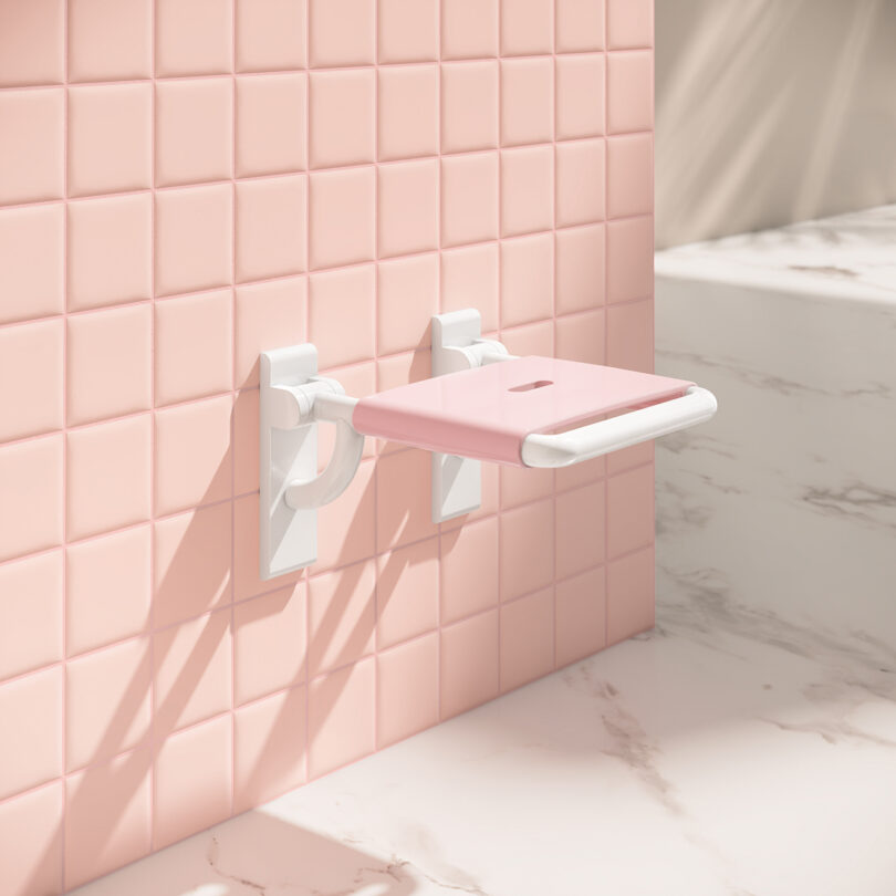 A pink foldable wall seat is mounted on pink square tiles next to a white marble surface in a bathroom.