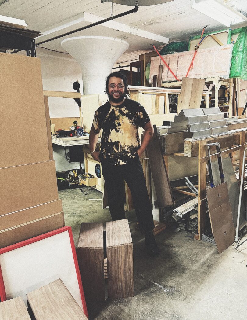 A person stands and smiles in a woodworking workshop, surrounded by wooden boards, tools, and unfinished furniture pieces