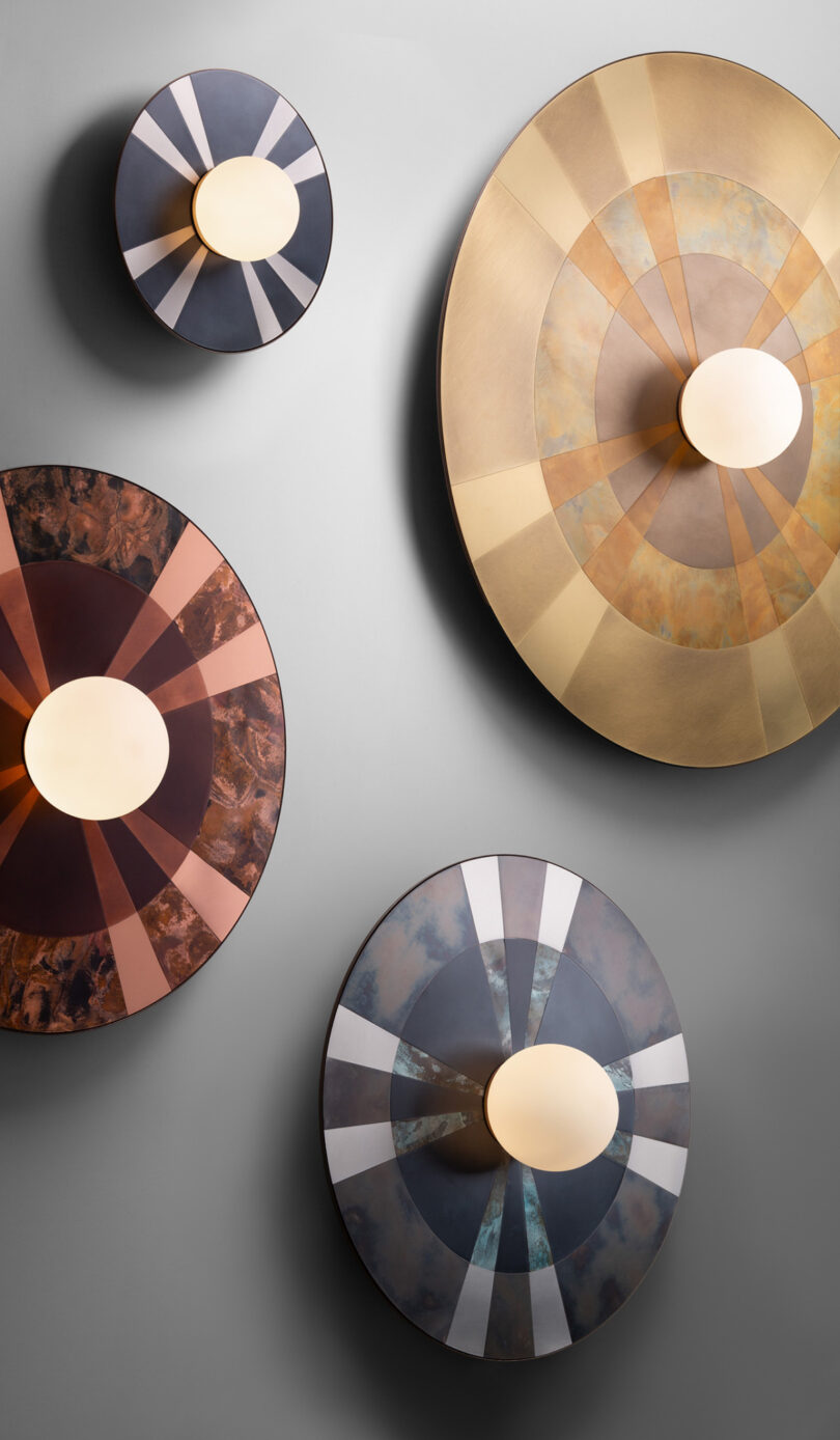 Four circular wall lights with geometric patterns and central light sources in various metal finishes on a gray wall.