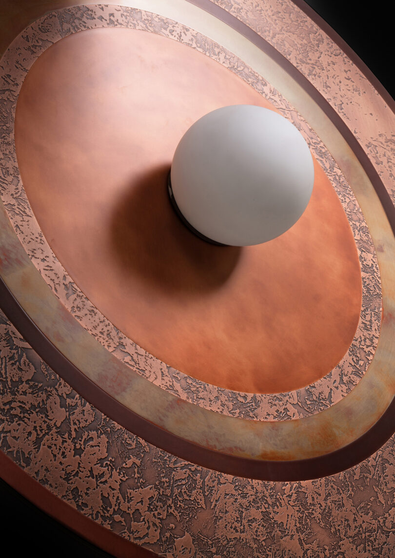 Close-up of a round, textured metallic light fixture with a central white globe light. The background is a dark, smooth surface.