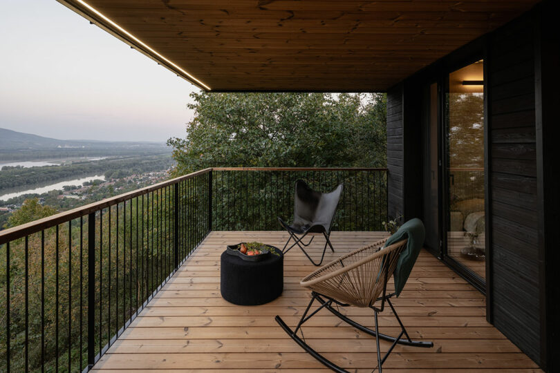 Wooden balcony with two chairs and a small table overlooks a scenic valley and river, surrounded by trees under a covered roof.