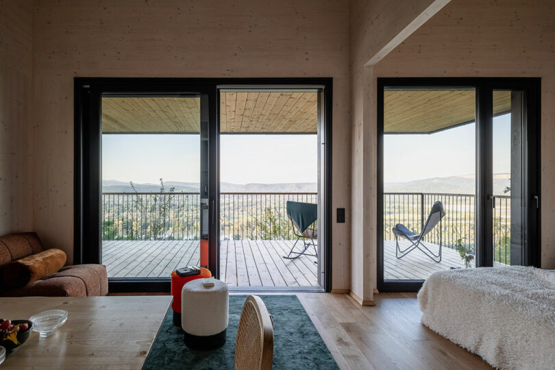 A modern room with wooden floors and large glass doors opens to a covered balcony with chairs, overlooking hills and a distant landscape.