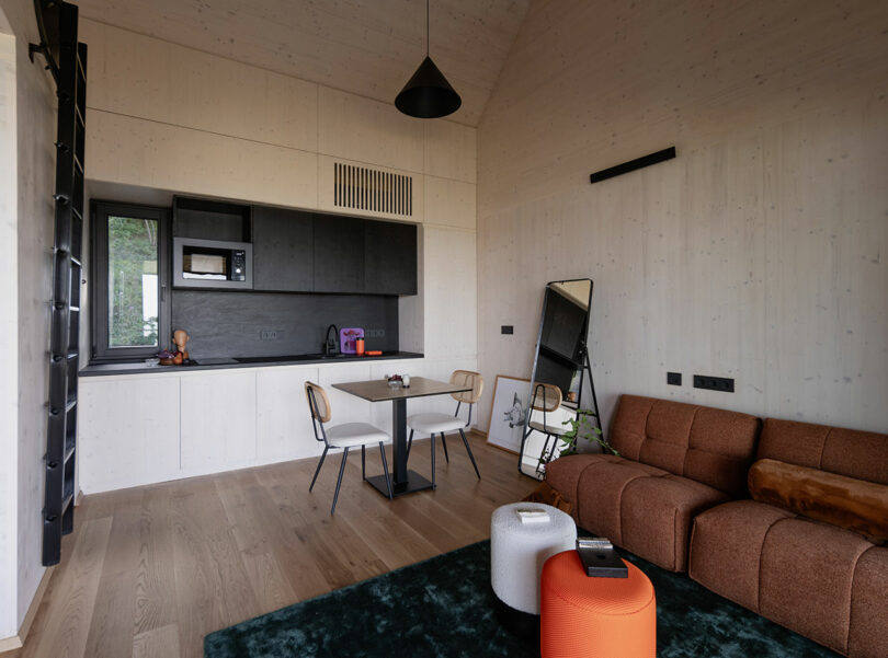 Modern studio apartment with a compact kitchenette, dining table, brown sofa, mirror, and contemporary decor in neutral tones.