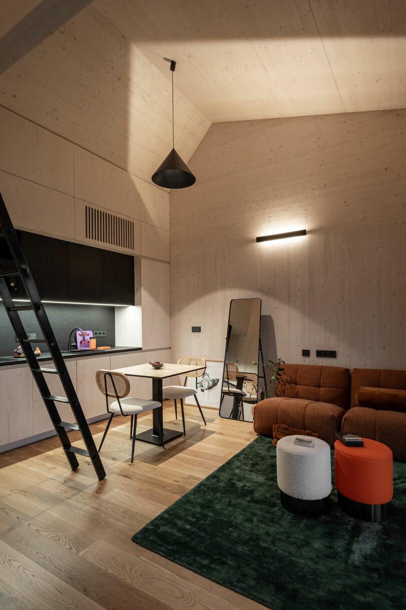 A modern living space with wooden walls, a brown sofa, a green rug, a black ladder, a dining table with two chairs, and a kitchenette under soft lighting.