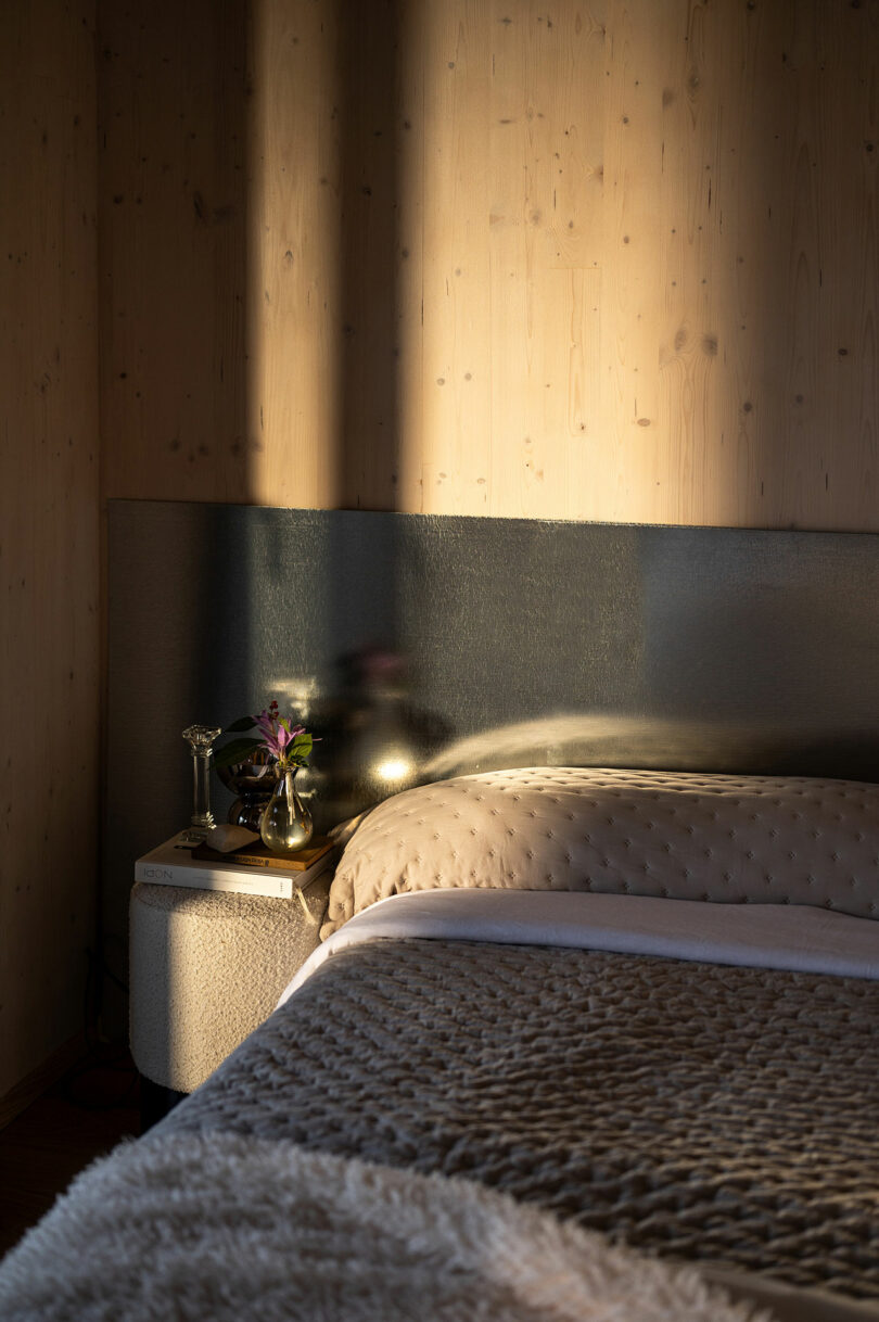 A neatly made bed with textured blankets sits beside a small nightstand holding a vase of flowers and a lamp, bathed in warm sunlight.