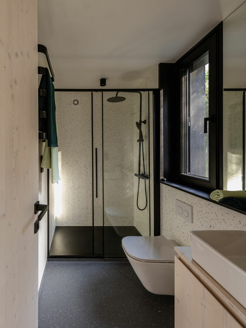 Modern bathroom with a glass shower enclosure, black fixtures, wall-mounted toilet, vessel sink, and large window letting in natural light.