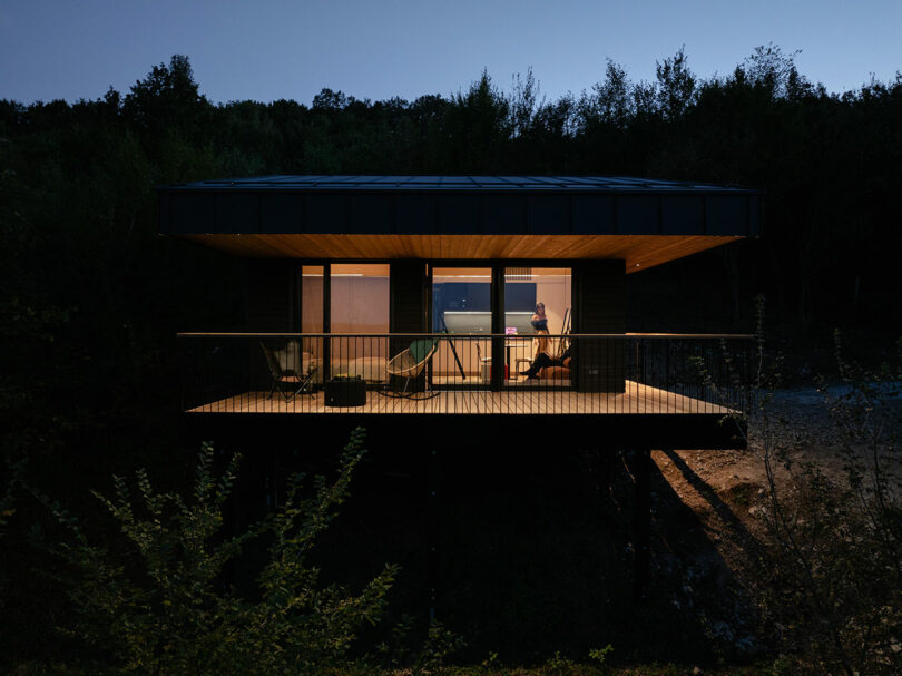 A modern house with large windows is lit from within at dusk, showing a person sitting at a table inside; the porch has chairs and is surrounded by trees.