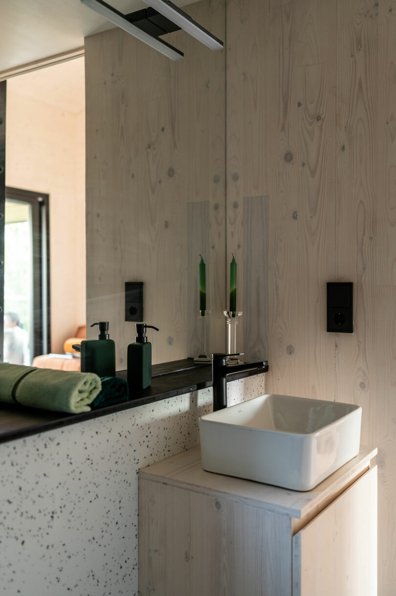 Modern bathroom with light wood walls, a white rectangular sink, green soap dispensers, green candles, and a folded green towel on a speckled countertop.