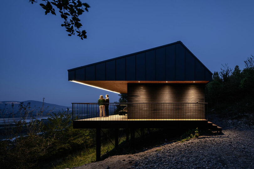A modern house with a large balcony on stilts is illuminated at dusk; two people stand on the balcony overlooking a scenic landscape.