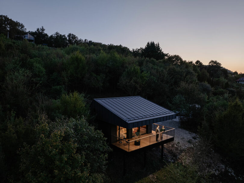 A modern cabin with a metal roof and large windows stands elevated on stilts among dense trees at dusk, with two people on the illuminated balcony.