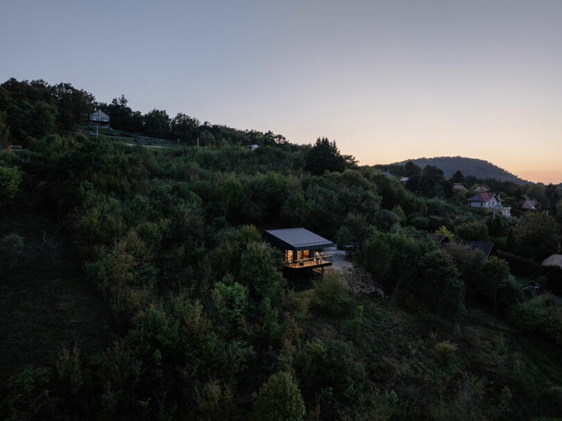 A small modern house with illuminated windows is surrounded by dense trees on a hillside at sunset, with other houses visible in the distance.