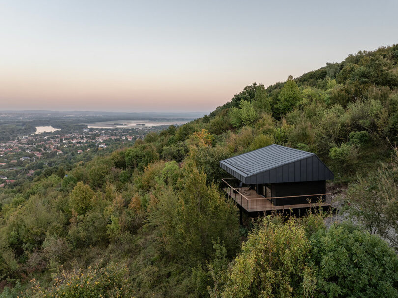 A modern black-roofed house sits on a forested hillside overlooking a valley with a river and a distant town under a clear sky.