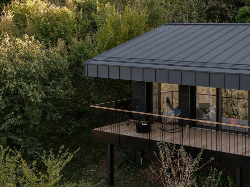 Modern black metal cabin with a covered balcony on stilts, surrounded by dense green trees and vegetation.