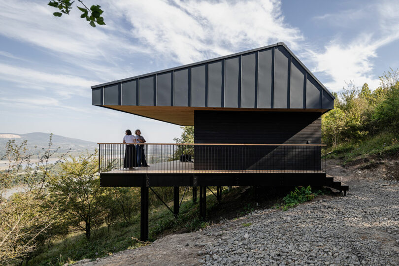 Modern black house on elevated stilts with a slanted roof, surrounded by trees; two people stand on the balcony overlooking a scenic landscape.